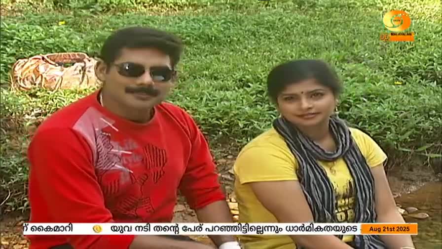 A man in sunglasses and a red shirt sits beside a woman in yellow, both facing the camera for a DD Malayalam broadcast. The bottom of the screen displays text in Malayalam.

