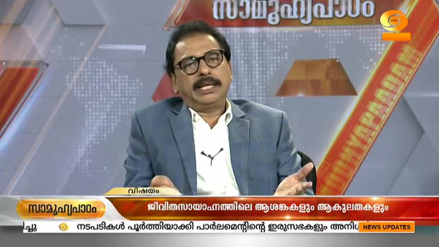 A man in a blue blazer and glasses gestures with his hands. He's speaking on a DD Malayalam news program, the title "Samuhyapaadam" visible above him.
