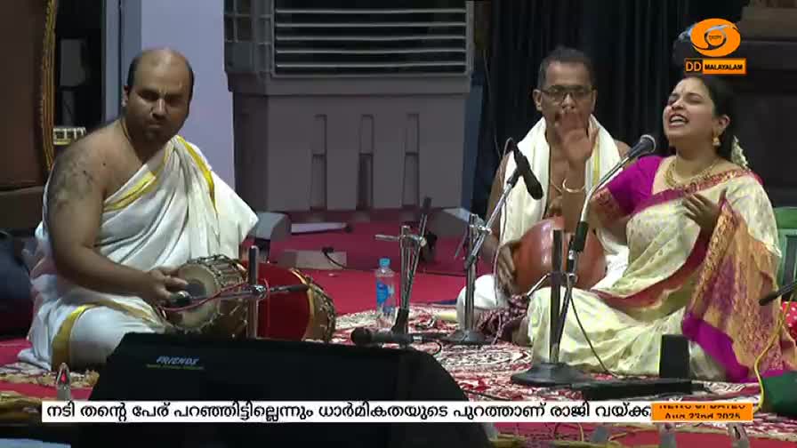 A woman in a sari sings into a microphone, her face lifted.  Two men accompany her, one on a hand drum and the other on a stringed instrument, all part of a performance on DD Malayalam.

