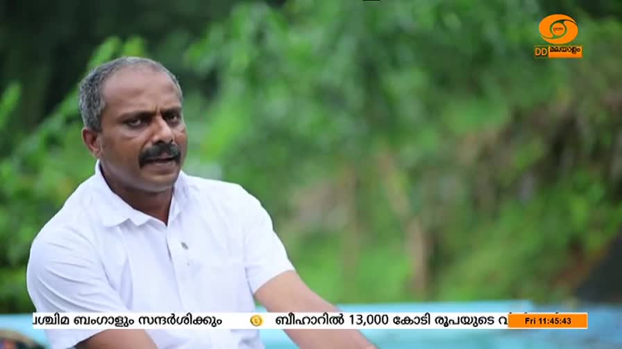 The man in the white shirt is speaking, his expression serious. DD Malayalam's logo is visible in the upper right corner, while text scrolls across the bottom of the screen.
