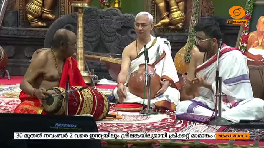 A man in a red and gold garment is playing a percussion instrument, his hands moving rhythmically. Two other men, one holding a pot, are seated nearby, likely part of a musical performance broadcast by DD Malayalam.
