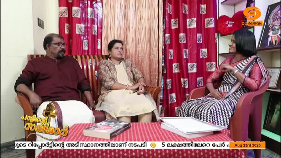 A woman in a black and white patterned saree sits across from a couple, all of them facing the camera. The DD Malayalam logo is visible in the corner, and a table with books sits in front of them.
