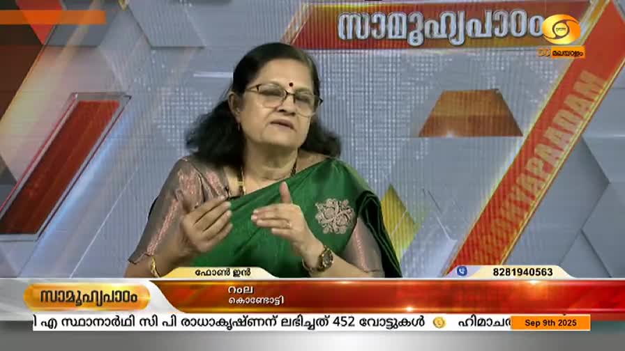 A woman in a green sari gestures with her hands while speaking, her glasses reflecting the studio lights. The DD Malayalam logo and text in Malayalam are visible on the screen behind her.
