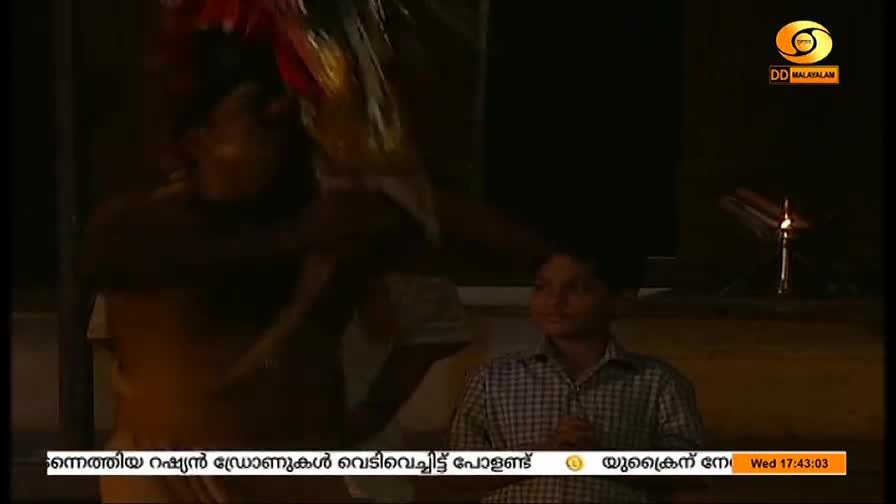 A person, obscured by movement, holds up a collection of colorful objects. A young boy in a plaid shirt watches, while the DD Malayalam logo and a news ticker are visible in the corner.
