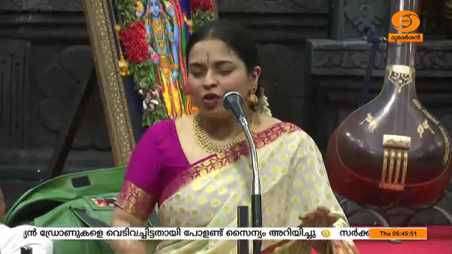 A woman in a vibrant sari sings into a microphone, her eyes closed, as a tanpura sits to her right. The DD Malayalam logo is visible, and the time is displayed in the corner.
