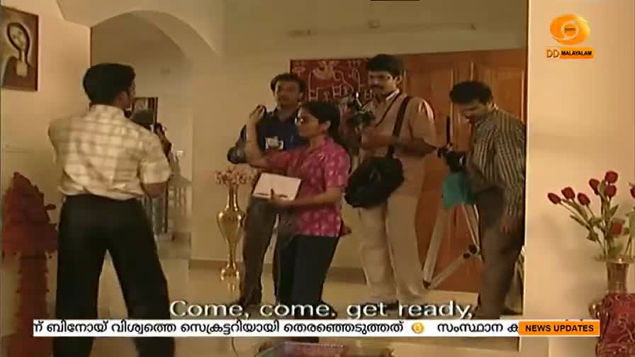 A group of people with cameras and microphones are preparing to film. A woman in a pink shirt holds a notebook, seemingly directing the scene for DD Malayalam.
