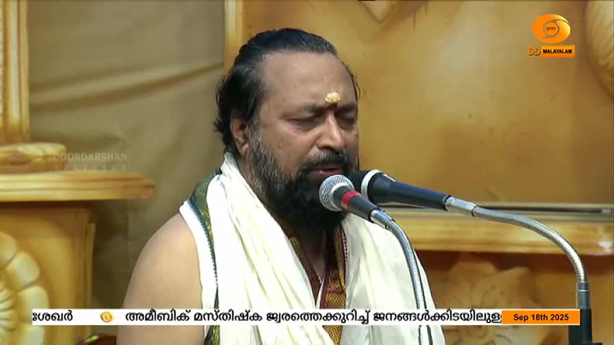 A man with a full beard and a white shawl speaks into a microphone, his eyes closed. The DD Malayalam logo is visible in the corner, and a date is displayed on the screen.

