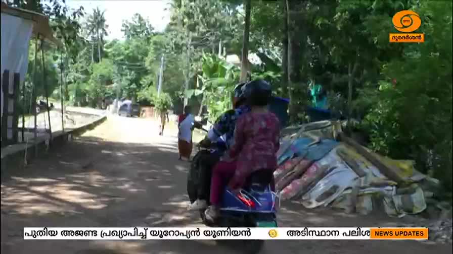A scooter, carrying two passengers in helmets, putters down a dirt road.  People are walking along the side, and the DD Malayalam logo is visible in the corner.
