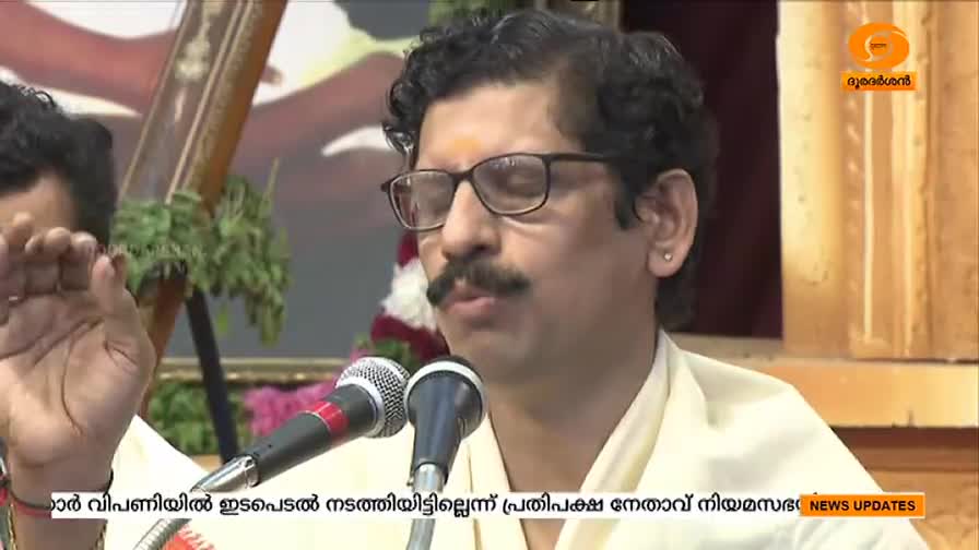 A man with glasses and a mustache speaks into a microphone, his hand gesturing as he talks. The DD Malayalam logo is visible in the corner.
