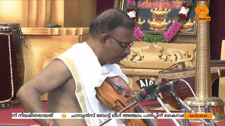A man in glasses, draped in a white cloth, intently plays the violin. The bow moves across the strings, and the music fills the space, likely broadcast on DD Malayalam.
