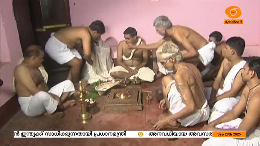 Several men, dressed in white dhotis, are gathered around a small fire, some attending to the ritual. One man is offering something to another, while others watch, all within a room that appears to be part of a ceremony.
