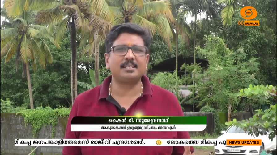 A man with glasses and a red shirt is speaking, likely for DD Malayalam. Behind him, a car sits parked near a lush green backdrop.
