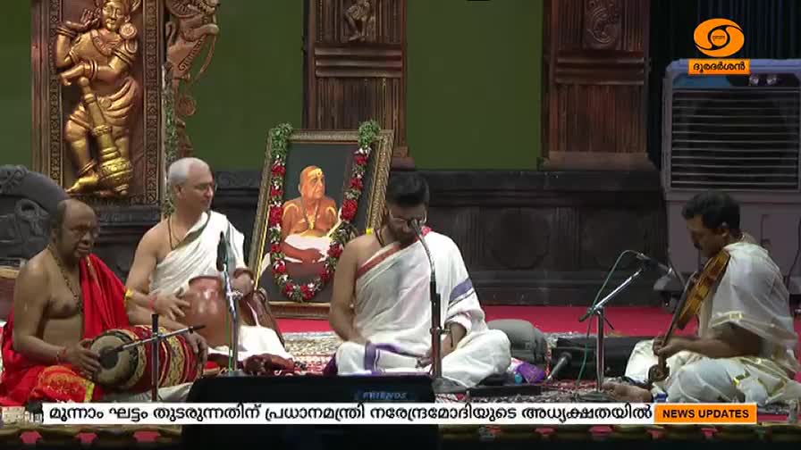 A group of musicians, dressed in white and red, are seated on the floor, performing. The DD Malayalam logo is visible in the corner.

