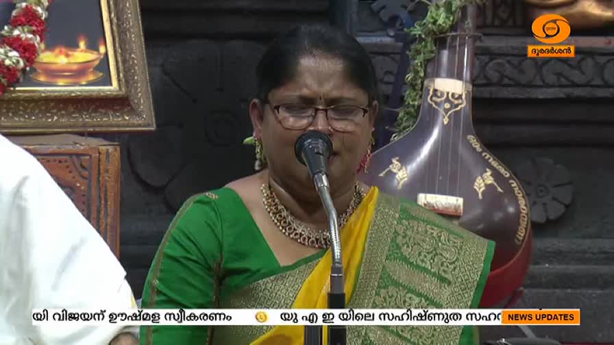 A woman in a green and gold sari sings into a microphone, her face illuminated by the light. Behind her, a large stringed instrument rests, and the DD Malayalam logo is visible in the corner.
