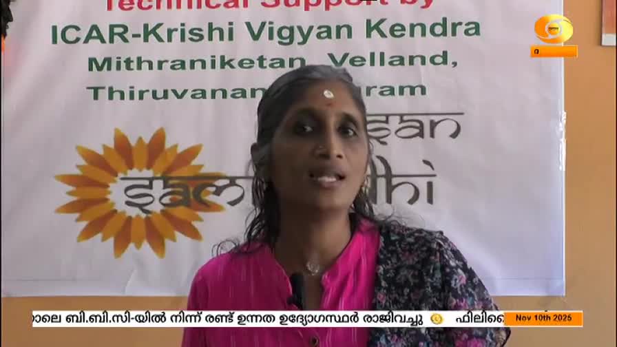 A woman in a pink top is speaking, her face framed by dark hair and a small bindi. Behind her, a banner with Malayalam text and a sunflower graphic is visible on what looks like a DD Malayalam broadcast.
