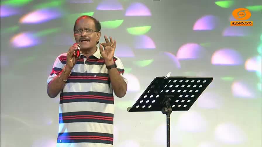 The man in the striped shirt holds a microphone, gesturing with his right hand as he speaks. Behind him, the stage lights cast a soft, colorful glow.
