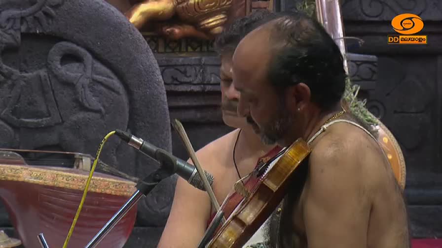 A man in India, likely performing, draws the bow across the violin strings. The instrument is held close to his body, and a microphone stands nearby.
