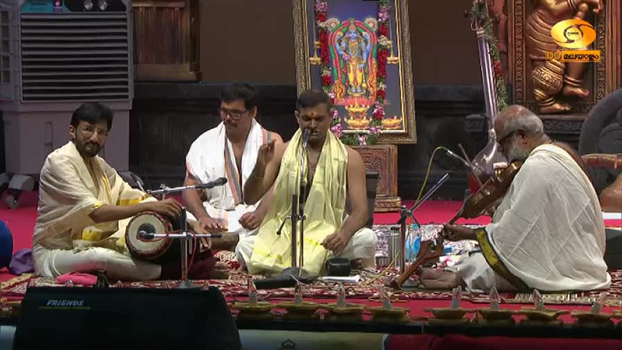 The vocalist, draped in a yellow shawl, gestures while singing into a microphone. A musician with a white beard draws his bow across a violin, adding to the music.
