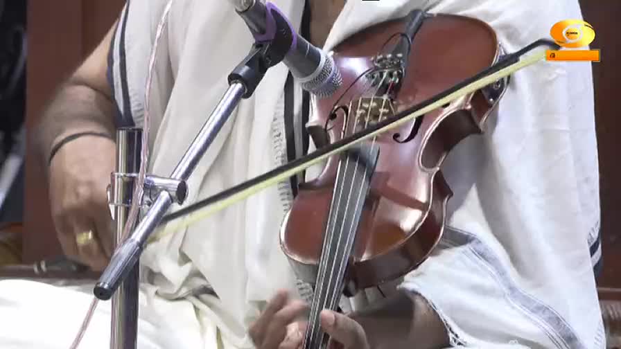 A musician, dressed in white, draws the bow across the violin's strings. The instrument is amplified by a microphone, likely for a broadcast on DD Malayalam.
A musician, dressed in white, draws the bow across the violin's strings. The instrument is amplified by a microphone, likely for a broadcast on DD Malayalam.