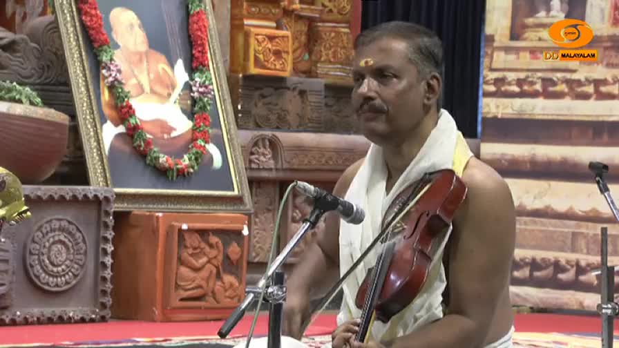 A man in India, on DD Malayalam, is playing the violin, his bow moving across the strings. A framed picture of a man with a garland hangs nearby.
A man in India, on DD Malayalam, is playing the violin, his bow moving across the strings. A framed picture of a man with a garland hangs nearby.