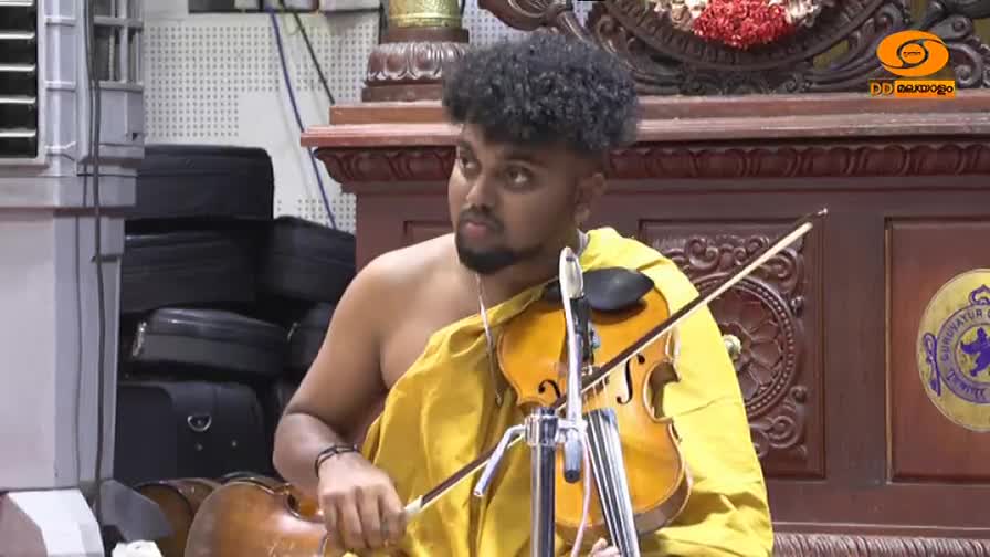 A young man, draped in a yellow cloth, holds a violin, the bow poised above the strings. He's focused, ready to play, as the DD Malayalam logo sits in the corner.
A young man, draped in a yellow cloth, holds a violin, the bow poised above the strings. He's focused, ready to play, as the DD Malayalam logo sits in the corner.
