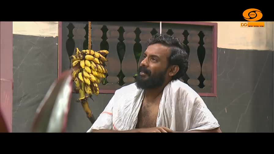 A man with a beard, wearing a white cloth, is speaking, his gaze directed upwards. A bunch of yellow bananas hangs from a rope nearby.
A man with a beard, wearing a white cloth, is speaking, his gaze directed upwards. A bunch of yellow bananas hangs from a rope nearby.