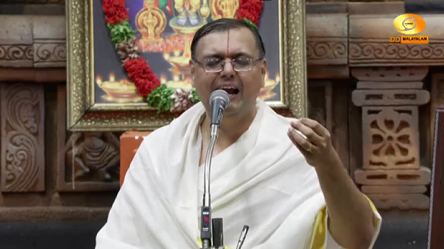 A man in glasses, wearing a white shawl, is singing into a microphone. His right hand is raised, and behind him is a framed image, all broadcast on DD Malayalam from India.
A man in glasses, wearing a white shawl, is singing into a microphone. His right hand is raised, and behind him is a framed image, all broadcast on DD Malayalam from India.