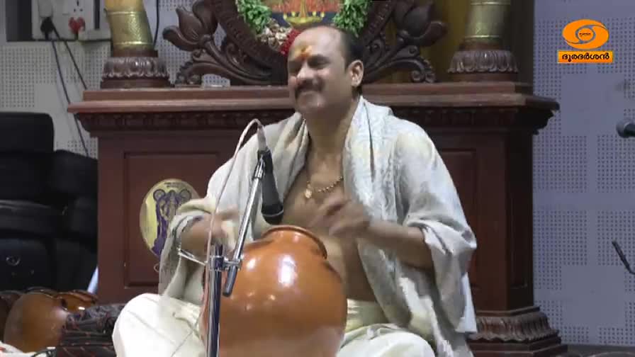 A man sits cross-legged, singing into a microphone with a clay pot drum in front of him. His hands move rhythmically as he plays, part of a performance on DD Malayalam.
A man sits cross-legged, singing into a microphone with a clay pot drum in front of him. His hands move rhythmically as he plays, part of a performance on DD Malayalam.