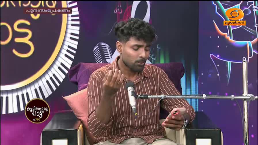 A young man, dressed in a striped shirt, sits on a plush chair, his gaze fixed on his phone. He gestures with his left hand as he sings into a microphone, his voice filling the studio. The backdrop features a large graphic of piano keys and the DD Malayalam logo. A young man, dressed in a striped shirt, sits on a plush chair, his gaze fixed on his phone. He gestures with his left hand as he sings into a microphone, his voice filling the studio. The backdrop features a large graphic of piano keys and the DD Malayalam logo.