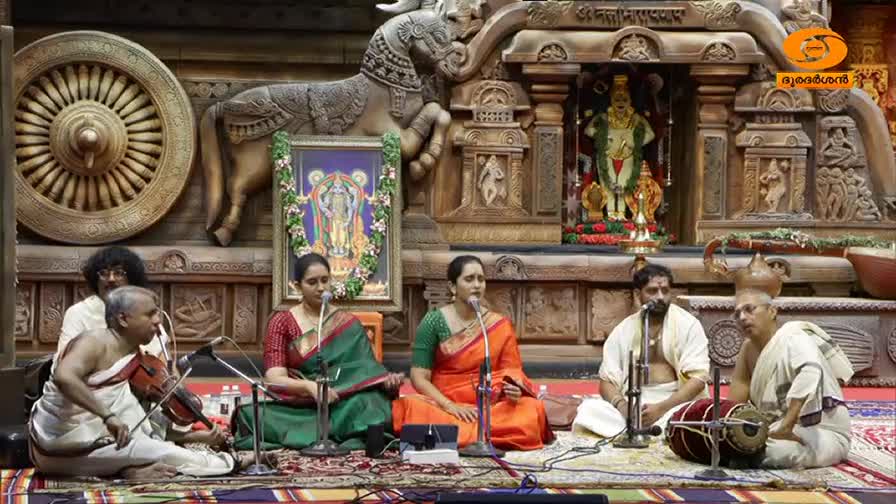 A violinist plays with his bow moving across the strings while two women sing into microphones, accompanied by a mridangam player and another musician. The performance unfolds against a backdrop of intricate wooden carvings and a framed deity, likely for a DD Malayalam broadcast.