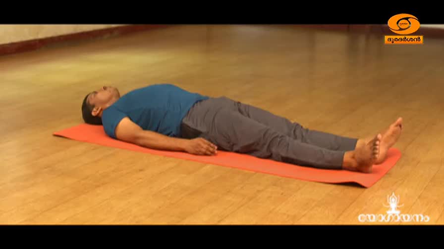 A man lies still on an orange mat, his arms resting by his sides. He's on a wooden floor, likely in a studio for a DD Malayalam program.