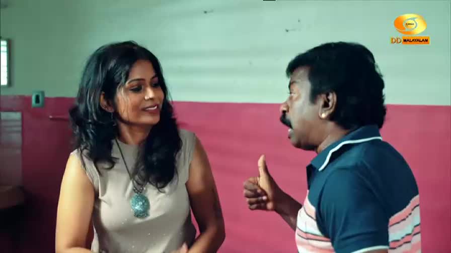 A man with a mustache gestures with his thumb up, speaking animatedly to a woman. The DD Malayalam logo is visible in the background. A man with a mustache gestures with his thumb up, speaking animatedly to a woman. The DD Malayalam logo is visible in the background.