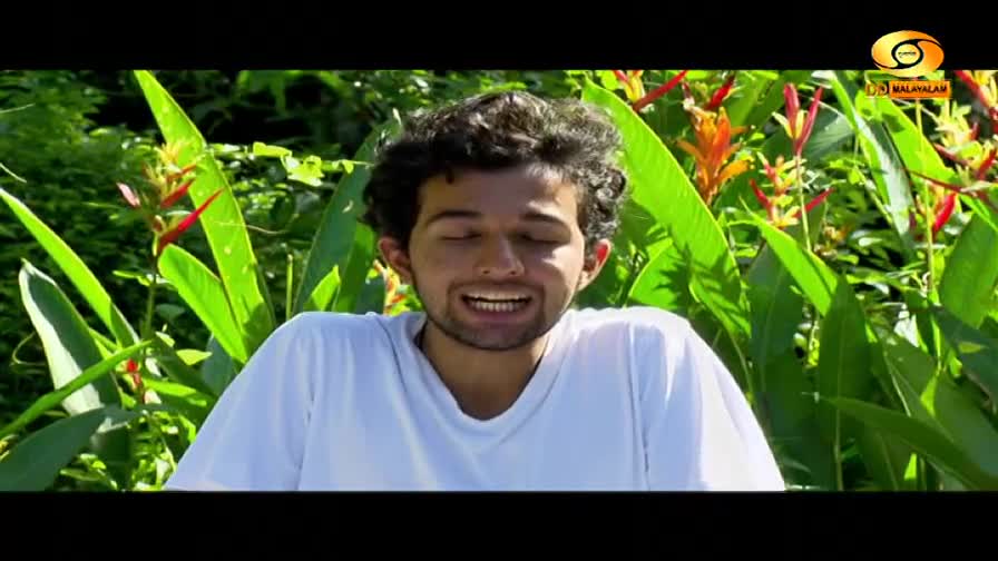 A young man with curly hair smiles broadly, his eyes closed, against a backdrop of lush green leaves and vibrant orange and red flowers. The DD Malayalam logo is visible in the upper right corner. A young man with curly hair smiles broadly, his eyes closed, against a backdrop of lush green leaves and vibrant orange and red flowers. The DD Malayalam logo is visible in the upper right corner.
