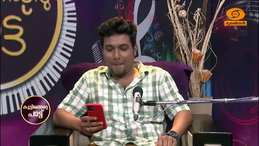 A young man in a checkered shirt looks down at his phone, a microphone positioned before him. Behind him, a large circular graphic with piano keys and Malayalam script hints at a music program on DD Malayalam. A young man in a checkered shirt looks down at his phone, a microphone positioned before him. Behind him, a large circular graphic with piano keys and Malayalam script hints at a music program on DD Malayalam.