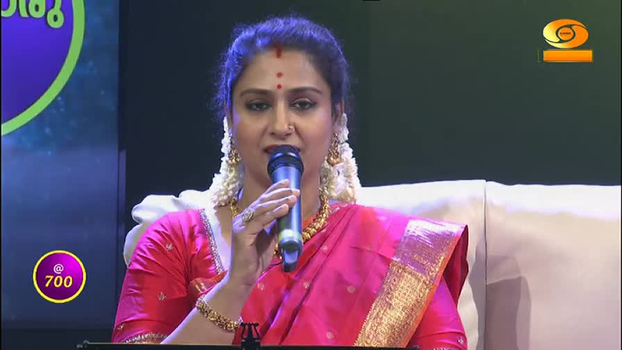 A woman in a bright pink sari speaks into a microphone, her bangles catching the light. The DD Malayalam logo is visible in the corner, suggesting a broadcast from India. A woman in a bright pink sari speaks into a microphone, her bangles catching the light. The DD Malayalam logo is visible in the corner, suggesting a broadcast from India.