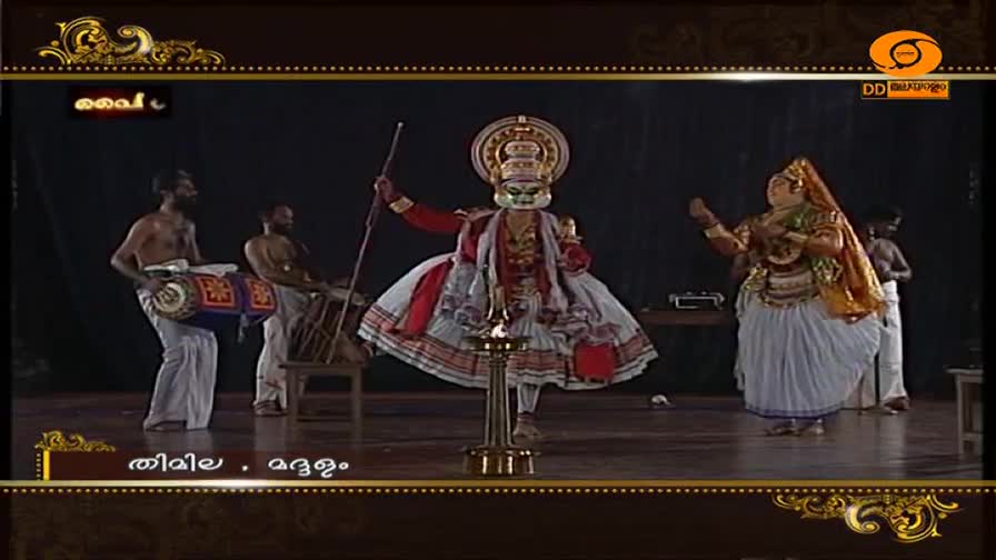 A Kathakali dancer with a striking green mask and elaborate costume stands center stage, holding a staff. To his left, two men play drums, their rhythmic beat filling the air. A Kathakali dancer with a striking green mask and elaborate costume stands center stage, holding a staff. To his left, two men play drums, their rhythmic beat filling the air.