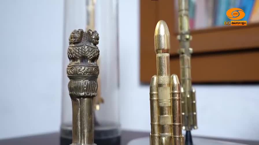 A brass Ashoka pillar emblem stands to the left, while to its right, a model of a rocket, constructed from what appears to be bullet casings, is displayed. The DD Malayalam logo is visible in the background. A brass Ashoka pillar emblem stands to the left, while to its right, a model of a rocket, constructed from what appears to be bullet casings, is displayed. The DD Malayalam logo is visible in the background.