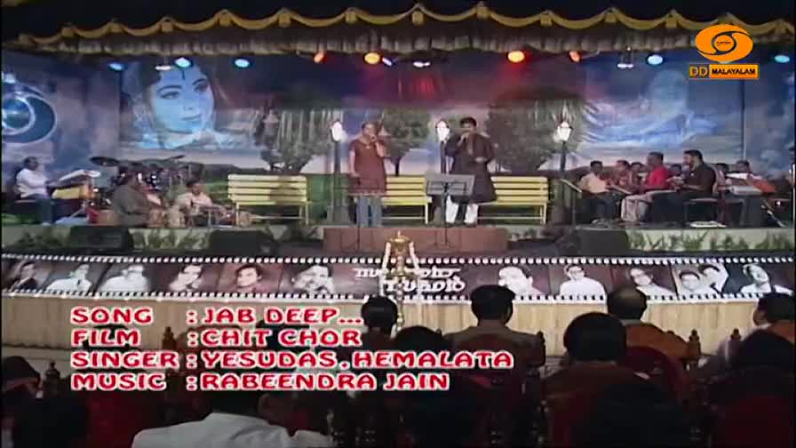 Two men stand on a stage, one in a maroon kurta and the other in a dark sherwani, singing into microphones. Behind them, a large portrait of a woman's face is projected onto a blue backdrop, and a band plays on benches. Two men stand on a stage, one in a maroon kurta and the other in a dark sherwani, singing into microphones. Behind them, a large portrait of a woman's face is projected onto a blue backdrop, and a band plays on benches.