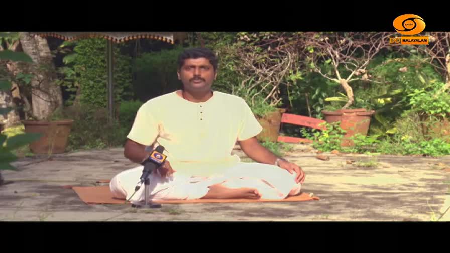 A man in a yellow kurta and white dhoti sits cross-legged on a mat, facing a microphone. Lush greenery and terracotta pots surround him in this outdoor setting. A man in a yellow kurta and white dhoti sits cross-legged on a mat, facing a microphone. Lush greenery and terracotta pots surround him in this outdoor setting.