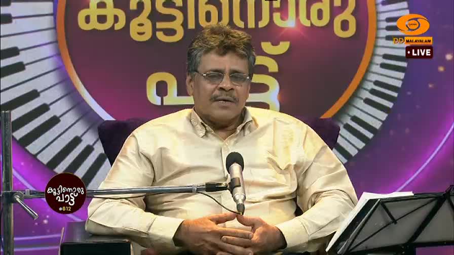 A man in a light gold shirt sits before a microphone on a stage, his hands clasped. Behind him, a large graphic displays piano keys and Malayalam text, with the DD Malayalam logo visible.