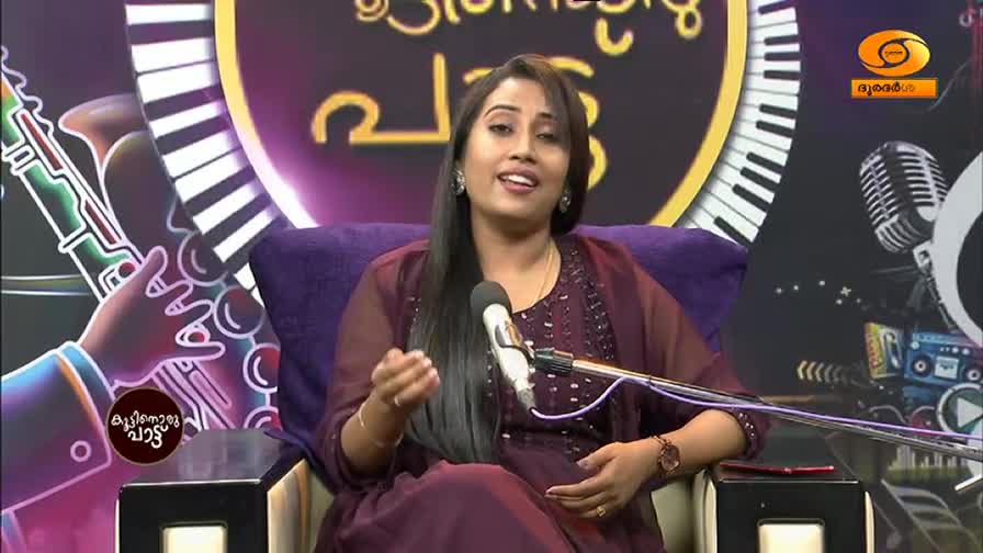 A woman in a deep purple dress speaks into a microphone, her hands gesturing animatedly. Behind her, a stylized keyboard and musical notes form part of the set for DD Malayalam.