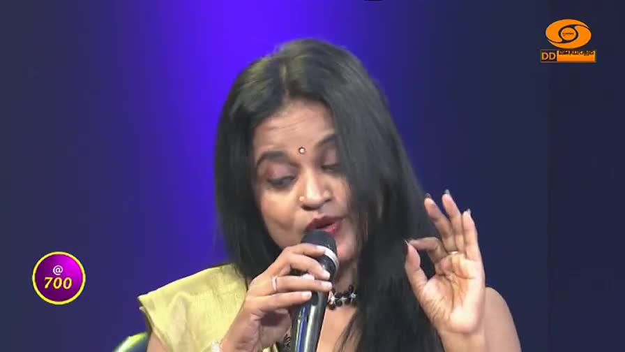 A woman sings into a microphone, her right hand raised with fingers forming a circle. The DD Malayalam logo is visible in the corner.