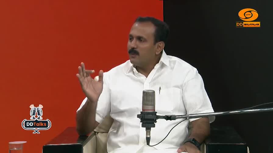 A man in a white shirt gestures with his left hand while speaking into a microphone. The DD Malayalam logo appears in the upper right corner.