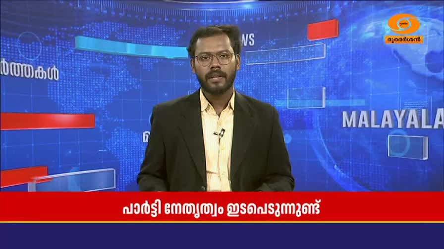 A man in a suit stands before a blue screen displaying a world map and the word "MALAYALAM." A red banner with Malayalam text runs across the bottom of the screen.