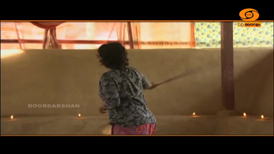 A person in a patterned shirt and pink skirt swings a long stick, its end a blur. Small diyas flicker along the low wall in front of them.