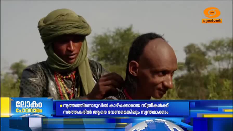 A man wearing a green turban and beaded necklaces adjusts the hair of another man. The DD Malayalam logo is visible in the upper right corner.