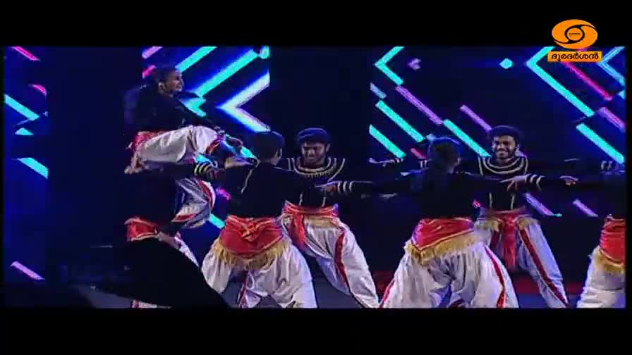 A group of dancers in traditional Kerala attire are performing on stage, their arms outstretched in unison. One dancer is lifted high above the others, a striking pose against the vibrant, geometric backdrop.