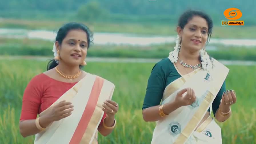 Two women in traditional white sarees with gold borders stand in a field, their hands gesturing as they sing. The DD Malayalam logo appears in the upper right corner.