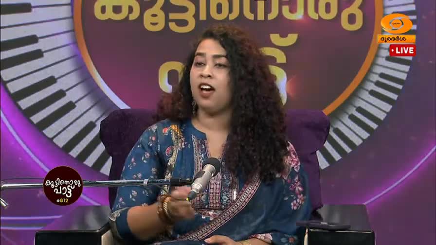 A woman with dark, curly hair is speaking into a microphone, her mouth open mid-sentence. She wears a blue kurta with a floral pattern, and a large keyboard graphic forms the backdrop of the set.