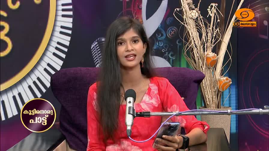 A young woman in a bright pink outfit sits on a purple chair, speaking into a microphone. She holds a smartphone in her right hand.